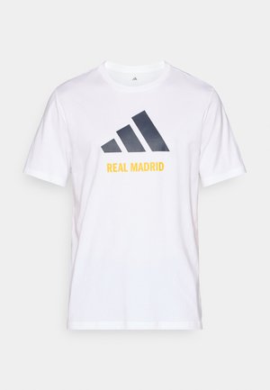 Balta medvilninė marškinėliai su navy blue Adidas logotipu ir "REAL MADRID" tekstu geltonai spausdintu po logotipu. Trumpi rankovės, apvalus kaklo dizainas.