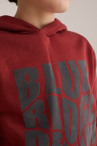 Raudona sportinė striukė su dideliu juodu piešiniu, kuriame užrašyti žodžiai "BLUE RIDGE". Medžiaga atrodo minkšta ir lygi; dizainas apima gobtuvą.