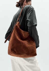 Hnědá semišová taška přes rameno s volnějším designem a dlouhým koženým popruhem, vystavená na světlém pozadí, se soft texturou.