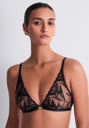 Femme portant un soutien-gorge en dentelle noire transparente avec motifs de feuilles et détail de fermeture dorée, cheveux tirés en arrière, expression neutre, fond uni gris clair.