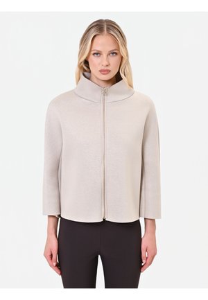Femme portant une veste beige clair zippée avec un col montant et des manches trois-quarts, associée à un pantalon noir, debout devant un fond blanc.