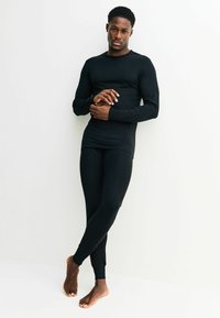 Fekete, hosszú ujjú felső és leggings sima anyagból, testhezálló szabás, minimális varratokkal és bokazáróval, egy álló modell viseli.