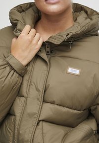 Olive green puffer jacket with a high collar, zip closure, elastic cuffs, and a logo patch on the front. Features quilted texture.