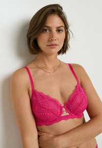 Una donna con i capelli corti castani indossa un reggiseno di pizzo rosa brillante e una delicata collana d'oro, posando su uno sfondo bianco.