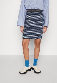 Jupe noire et blanche à pois, longueur genoux, avec une ceinture contrastante et des bandes latérales. Associée à des chaussettes bleues et des bottines blanches.