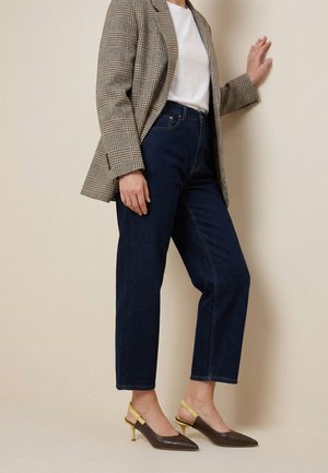 Persoon staat tegen een beige muur, draagt een bruine geruite blazer, een wit overhemd, donkerblauwe spijkerbroek en bruine puntige slingback hakken met gele bandjes.