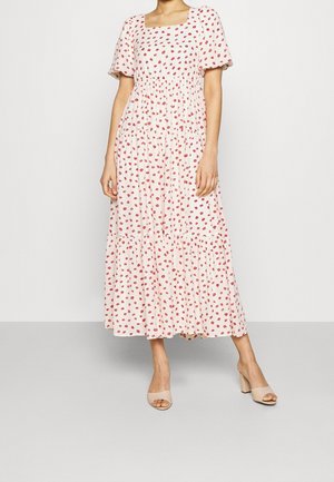 Robe maxi florale rose avec de courtes manches bouffantes, taille élastique, jupe à volants et petit motif de fleurs rouges partout. Portée avec des talons beiges.