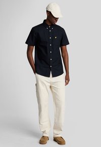 Camicetta blu marina a maniche corte con taschino e logo giallo, abbinata a pantaloni cargo crema e scarpe slip-on marroni chiari.