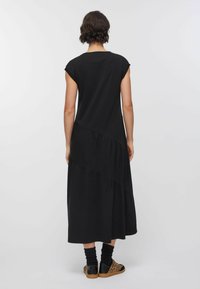 Robe maxi noire avec manches courtes en forme de pélerine, présentant un design à couches et une texture semi-transparente. Portée avec des baskets à motifs et des chaussettes noires.
