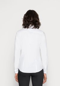 Person with shoulder-length dark hair wearing a fitted white long-sleeve shirt and black pants, shown from the back against a plain background.