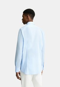 Camicia a maniche lunghe azzurro chiaro realizzata in tessuto morbido, con colletto classico, polsini con bottoni e orlo curvo. Vista da dietro.