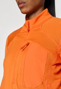 Oransje langermet topp med glidelåsåpning i halsen, teksturert stoff og en stor frontlomme med snorakksent.