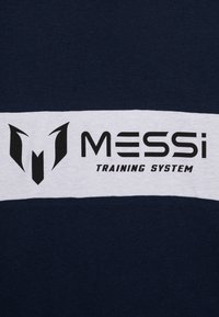 Marineblå T-skjorte med en hvit horisontal stripe som har svart "MESSI"-logo og teksten "TRAINING SYSTEM". Mykt bomullsmateriale.