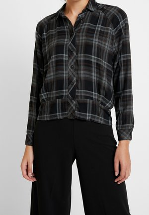 Kvinde iført en sort, grå og brun ternet skjorte med knapper og lange ærmer samt sorte bukser mod en ensartet lys baggrund.