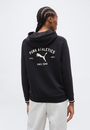 Femeie cu părul împletit, purtând un hanorac negru cu logo-ul și textul "Puma Athletics" și "Since 1948," asortat cu pantaloni sport gri deschis.