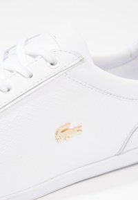 Bílá teniska s texturovaným koženým svrškem, plochými bílými tkaničkami a zlatým akcentem loga krokodýla na boku. Kožené detaily podél podrážky.