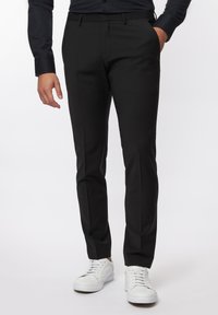 Schwarze, maßgeschneiderte Hose mit slim fit, mit flachem Frontteil, Seitentaschen und Gürtelschlaufen, kombiniert mit weißen Sneakern.