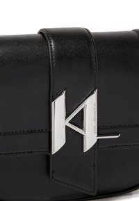 Una borsa a mano in pelle nera presenta una texture liscia, una forma strutturata e un accento del logo in argento sul davanti con dettagli di cucitura orizzontale.
