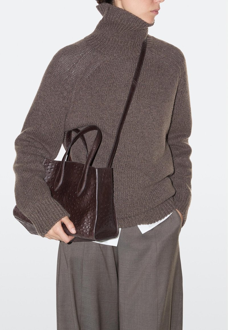 Persona che indossa un maglione a collo alto marrone spesso e pantaloni grigi, che tiene in mano una borsa a tracolla in pelle marrone scuro con doppie maniglie e tracolla.