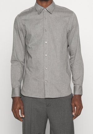 Mężczyzna ubrany w dopasowaną jasnoszarą, długorękawową koszulę na guziki z kołnierzykiem oraz pasujące szare spodnie, stoi z ramionami luźno opuszczonymi.