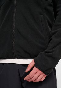 Svart fleecejacka med dragkedja fram, strukturerad tyg, och tumhål vid ärmsluten, med sidofickor och ett vitt innerlager.