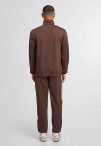 Brūns treniņtērps ar augstu apkakli un sānu svītrām biksēs. Veidots ar brīvu piegriezumu un mīkstu auduma tekstūru.