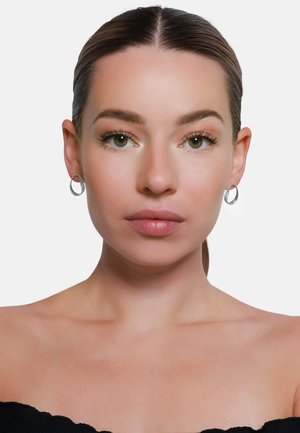 Orecchini a cerchio in argento presentano un design circolare e liscio, che riflette la luce. Il modello ha un trucco naturale e uno sfondo neutro.