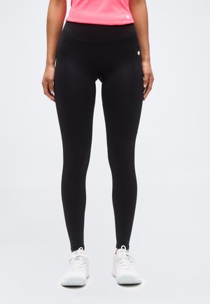 Persoon die zwarte legging met hoge taille en zijzak en witte sneakers draagt, staand tegen een effen lichte achtergrond.