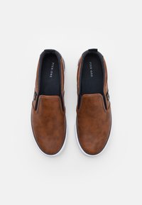 Braune Slipper aus Kunstleder mit glatter Textur, schwarzen elastischen Akzenten und weißen Gummisohlen. Markenname innen sichtbar.