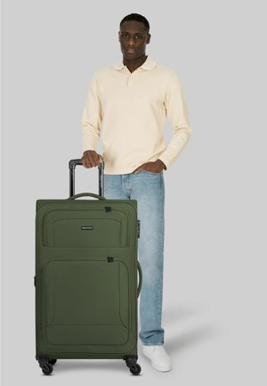 Man staat met zijn rechterhand op het verlengde handvat van een grote groene trolleykoffer, gekleed in een beige shirt met lange mouwen, blauwe spijkerbroek en witte schoenen.