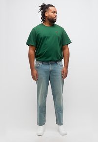 Άνδρας με κοτσίδες, φοράει πράσινο μπλουζάκι, ανοιχτό μπλε τζιν και άσπρα αθλητικά παπούτσια, στέκεται και κοιτάζει προς το πλάι μπροστά σε απλό φόντο.