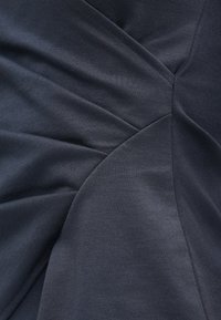 Tecido cinza escuro dobrado e costurado num padrão radial, criando seções curvas sobrepostas com linhas de costura visíveis.