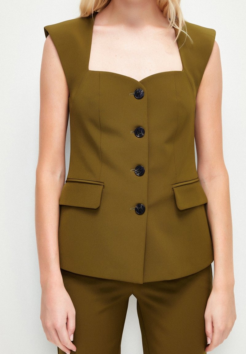 Femme portant un blazer sans manches vert olive avec des boutons noirs et un pantalon assorti, debout devant un fond blanc.