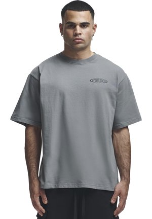 Férfi, aki laza szürke pólót visel, amelyen a "2Y STUDIOS" logó található a mellkasán, fehér háttér előtt, előre nézve áll.