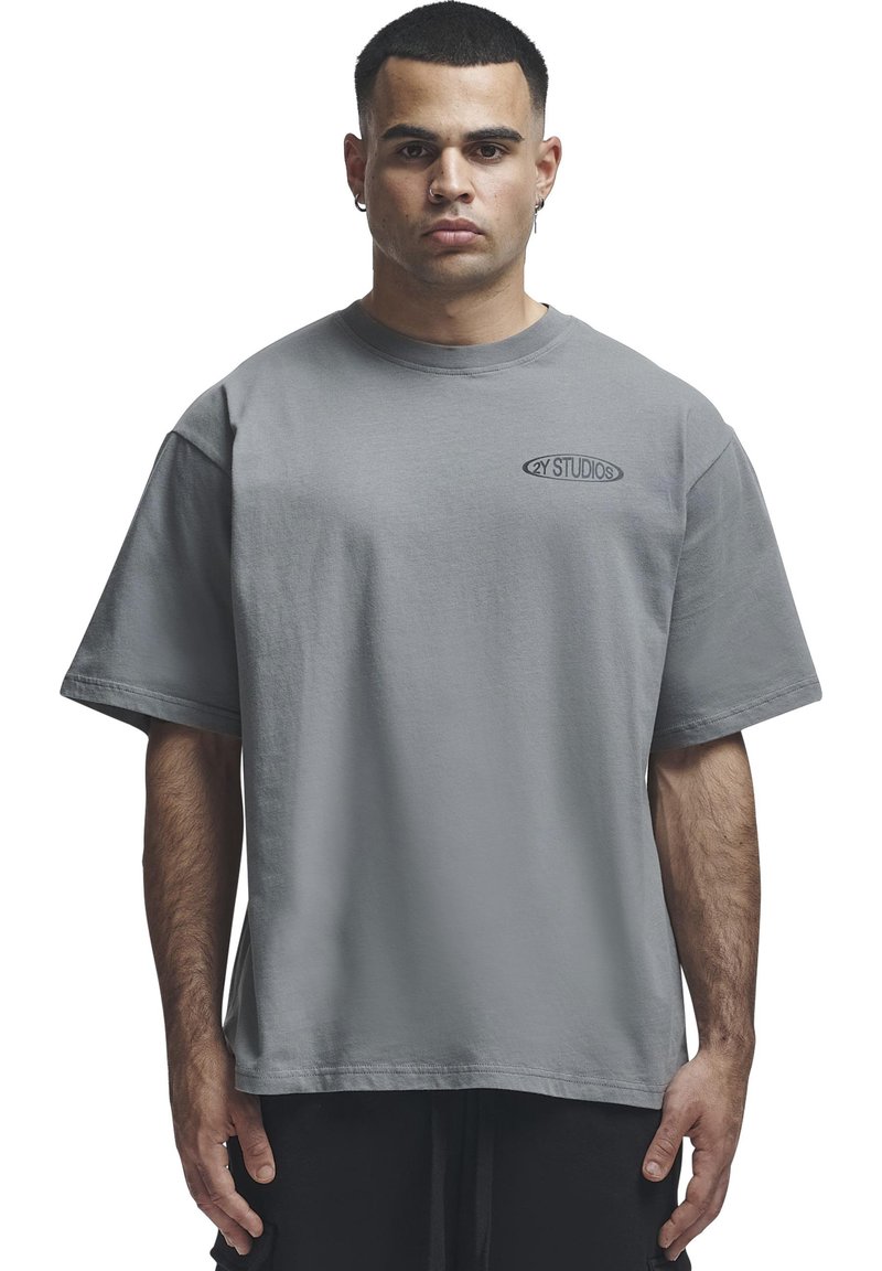 Homme portant un t-shirt gris ample avec le logo "2Y STUDIOS" sur la poitrine, debout face à un fond blanc.