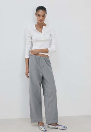 Mujer de pie frente a una pared blanca, vestida con una camisa blanca de manga larga, pantalones grises sueltos y zapatos planos azules y blancos con estampado.