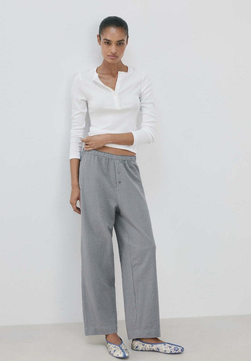 Femme debout contre un mur blanc, vêtue d'une chemise blanche à manches longues, d’un pantalon gris ample et de chaussures plates bleues et blanches à motifs.