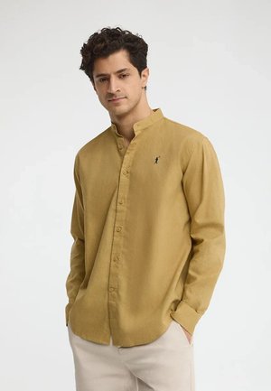 Uomo con capelli ricci scuri indossa una camicia senape a maniche lunghe con colletto alla coreana e pantaloni beige chiaro, mani nelle tasche.
