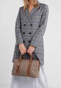 Brązowa torebka w wzory z podwójnymi zamkami błyskawicznymi i jasnobeżowymi skórzanymi uchwytami. Nosi ją osoba w szarym, w kratkę, podwójnym płaszczu na lekkiej spódnicy.