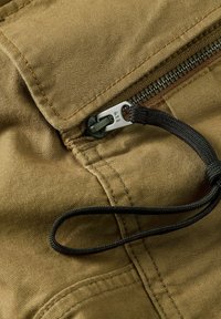 Beige canvasstof heeft een ritssluiting met een zilveren trekker gemarkeerd "RAW" en een zwarte koordbevestiging nabij de naad.