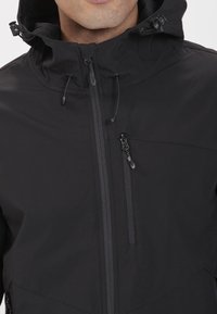 Giacca nera con zip, caratterizzata da un colletto rialzato, cappuccio con cordino e due tasche con zip. Il materiale è liscio con una finitura opaca.