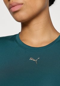 Szmaragdowa koszulka sportowa z okrągłym dekoltem, z metalicznym logo Pumy na klatce piersiowej. Materiał ma gładką, śliską fakturę.