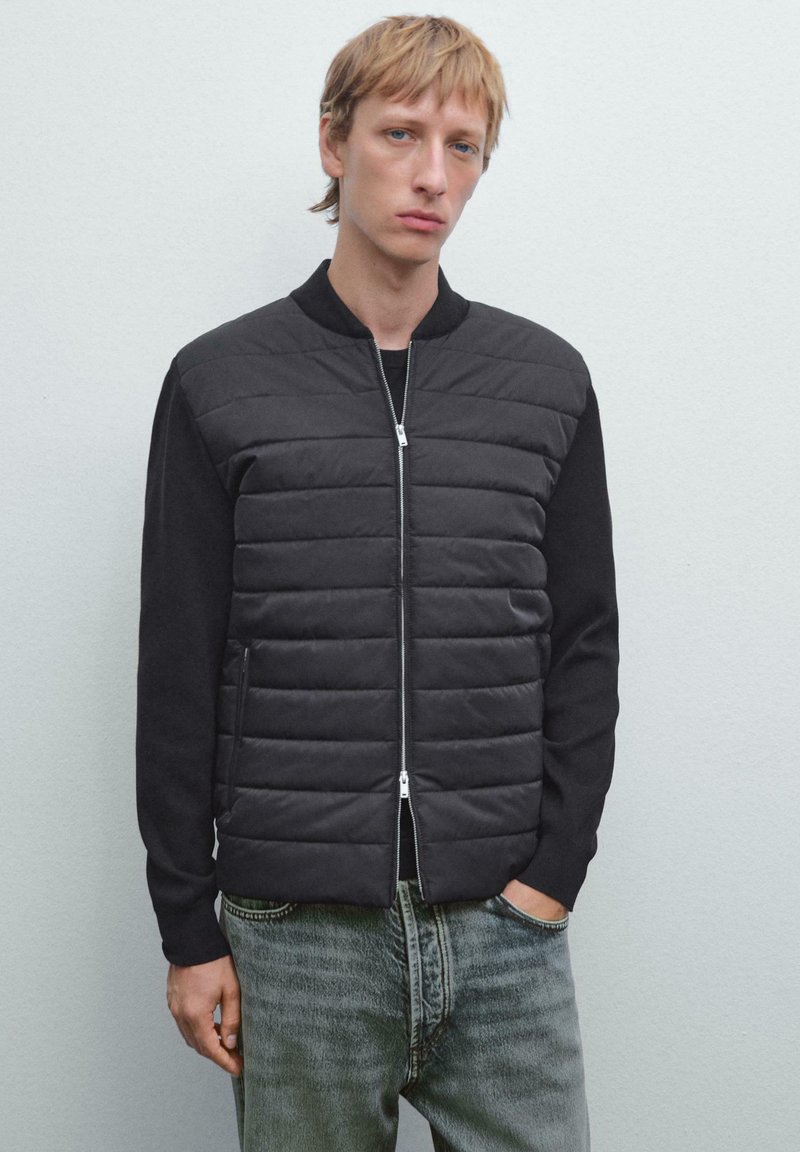 Giubbotto bomber nero con parte anteriore trapuntata, maniche a maglia e chiusura con zip; abbinato a jeans in denim sbiadito. Texture liscia e opaca.