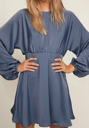 Kobieta ubrana w luźną, długorękawową, ciemnoniebieską sukienkę z marszczoną talią i elastycznymi mankietami, z rękami opartymi na biodrach na jasnym tle.