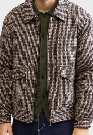Homme portant une veste à carreaux marron et beige sur un cardigan boutonné vert olive avec un jean bleu foncé, montré du cou à la taille.