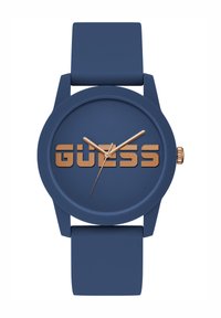 Kék szilikon karóra matt felülettel, kerek számlappal, aranyszínű mutatókkal és a középen aranyban dombornyomott „GUESS” logóval.