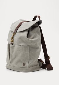 Mochila de lienzo gris claro con bolsillo frontal, correas ajustables marrones y cierre de clasp de latón sobre un fondo blanco sólido.
