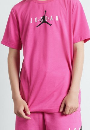 Dítě nosí jasně růžové tričko Jordan s černým logem Jumpman a odpovídající kraťasy, jednou rukou drží okraj trička.