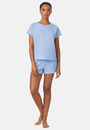 Mujer de pie descalza que lleva una camisa de manga corta a rayas azul claro y blanca y pantalones cortos a juego contra un fondo liso.