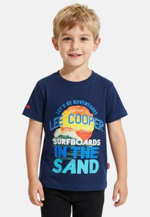 Jeune garçon aux cheveux blonds portant un t-shirt bleu marine avec un motif coloré de coucher de soleil et de planche de surf accompagné d'un texte.